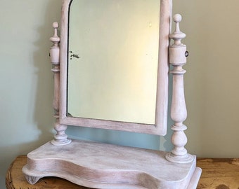 May include: A vintage-style vanity mirror with a light-colored, distressed finish. The arched mirror is supported by ornate, turned posts and a decorative base. The mirror is designed to sit on a table or dresser.