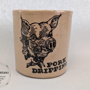 以下が含まれることがあります： 豚の頭の黒いイラストと「Pork Dripping」の文字が入った、ベージュ色の陶器の瓶。瓶は円筒形で、縁がわずかに広がっています。イラストは詳細で、ヴィンテージ風です。