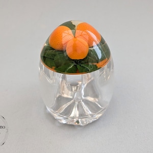 May include: A clear glass decorative object with a dome-shaped top. The top features several orange fruits and green leaves. The base of the object is clear and has a decorative design. The object is on a light grey surface.