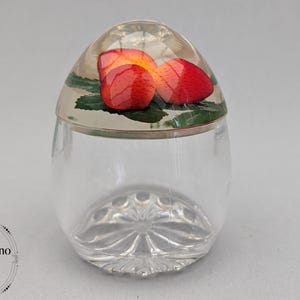 May include: A clear glass jar with a rounded lid featuring an embedded design of red strawberries and green leaves. The base of the jar has a pressed radial pattern. The text StudioVintano Vintage Treasures Curios, Designs & Gifts On Etsy is visible.