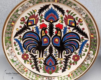 Peut inclure: Une assiette en céramique blanche avec un bord doré présente un motif coloré de deux coqs se faisant face. Les coqs sont noirs avec des crêtes et des barbillons rouges, des plumes bleues et rouges et des accents dorés. L'assiette est entourée d'une bordure verte et dorée avec un motif répétitif de volutes et de points. L'assiette est signée "Studio Vintano & Milano Bespoke Studio Collaboration."