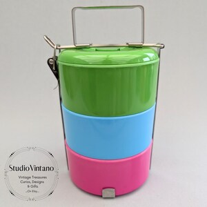 May include: A three-tiered lunch container in green, blue, and pink. It has a metal handle and clasps. The container is designed to stack, with a rectangular handle for carrying. Each section is a different bright colour.