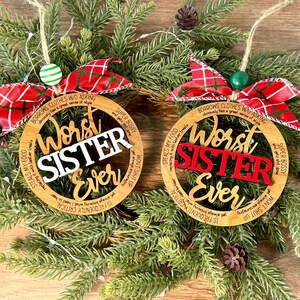 May include: Two wooden Christmas ornaments with the text "Worst Sister Ever". One ornament has white text, the other has red text. Both have red and white plaid bows and are surrounded by evergreen branches.