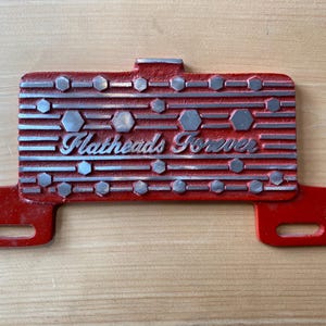 May include: A red metal sign with the words "Flatheads Forever" in silver script. The sign has a rectangular shape with horizontal lines and silver geometric shapes. It has two rectangular extensions with holes.