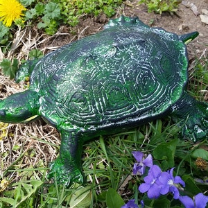 May include: A green turtle statue with a textured shell and detailed features. The statue is placed in a natural outdoor setting with grass, dandelions, and purple flowers, creating a garden ornament.