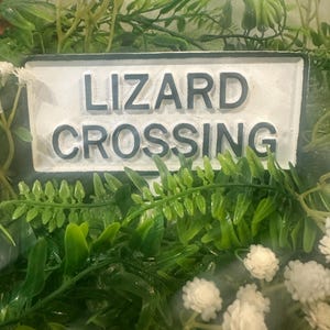 May include: A rectangular sign with the words "LIZARD CROSSING" in dark gray letters. The sign is surrounded by green foliage, including ferns and small white flowers. The sign has a white background.