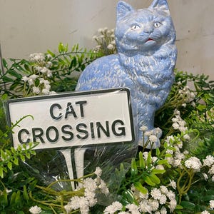 May include: A light blue cat figurine with textured fur and white highlights, positioned behind a rectangular white sign with a black border and the black text CAT CROSSING, surrounded by green foliage and small white flowers.