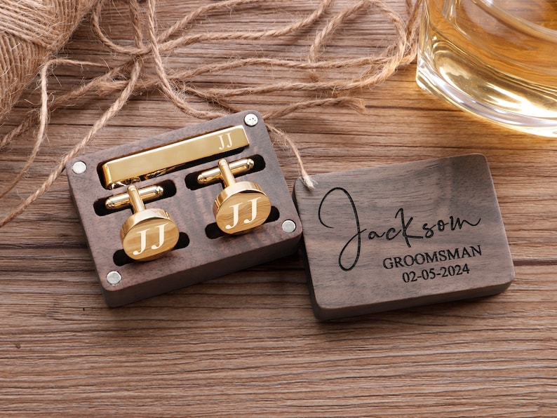 Puede incluir: Una caja de madera con un clip de corbata y gemelos de oro. La caja est&aacute; grabada con el nombre "Jackson" y las palabras "Groomsman 02-05-2024".
