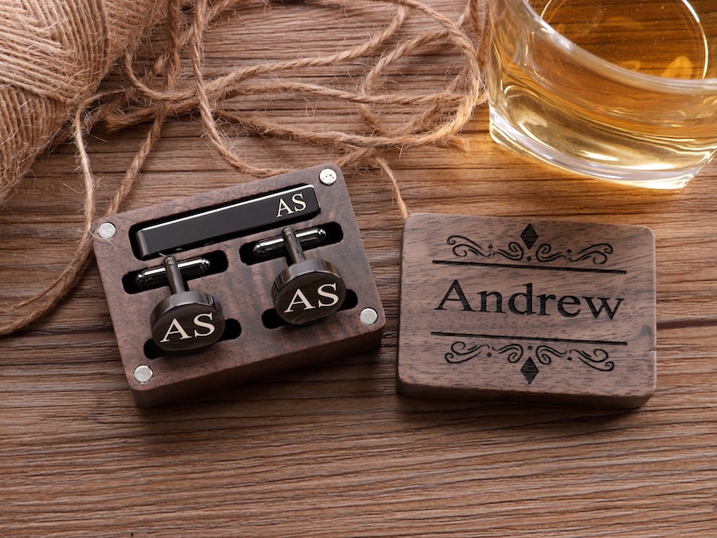 Puede incluir: Una caja de madera con un clip de corbata negro y gemelos. La caja est&aacute; grabada con las iniciales "AS" y el nombre "Andrew".