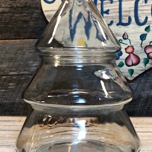Puede incluir: Un tarro de cristal transparente con forma de árbol de Navidad. El tarro tiene una tapa cónica y una base escalonada. El fondo incluye una superficie de madera y un corazón de madera decorativo con la palabra "WELCOME".