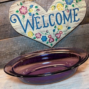 May include: A purple oval glass dish with a black rim. Behind the dish is a heart-shaped wooden sign with the word "WELCOME" in blue script, surrounded by painted flowers.