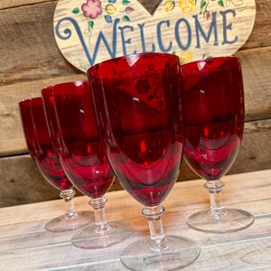 May include: A set of four deep red glass goblets with clear stems and bases. Each goblet features a smooth, rounded bowl. The base of the front goblet is etched with the text "ROBERT STANLEY HOME COLLECTION" in small, clear, capitalized letters.