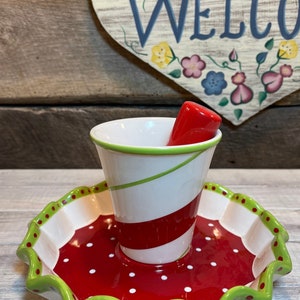 以下が含まれることがあります： 赤と白のストライプのセラミック製サービングディッシュで、緑色の縁取りと白い水玉模様が付いています。ディッシュには、緑色の縁取りが付いた赤と白のストライプのセラミックカップが付属しています。カップには赤いハンドルが付いています。