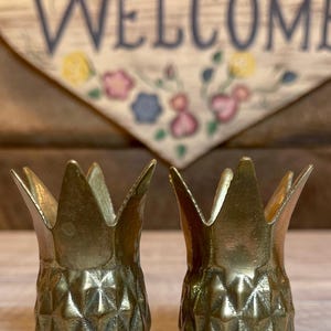 May include: A pair of metallic gold-toned pineapple-shaped vessels featuring a textured diamond-patterned body and a pointed, leaf-like rim. These two identical containers are crafted with a polished finish and stand upright as decorative objects.
