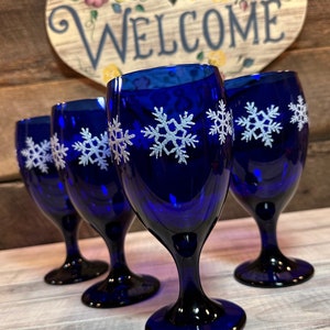 May include: Four cobalt blue wine glasses with white snowflake designs. The glasses are arranged in a semi-circle on a light wood surface.