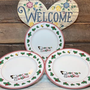 May include: Three white ceramic plates with a red border, holly and berry design, and the phrase "Joy to the World" with a Snoopy graphic. A wooden heart-shaped "Welcome" sign is in the background.