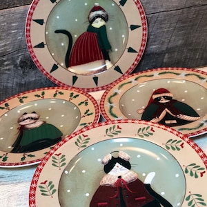 May include: Set of four decorative plates featuring whimsical cat illustrations in winter attire. Each plate has a beige border with red and green accents, and a central image of a cat wearing a hat and coat against a snowy background.