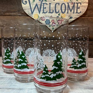 May include: Four clear drinking glasses with a winter scene design. The glasses feature green pine trees, white trees, and falling snow. A red stripe encircles the base of each glass. A wooden heart-shaped sign with the word "WELCOME" is in the background.