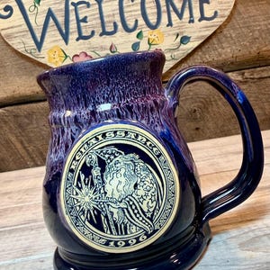 May include: A deep blue ceramic mug with a glossy finish and a lighter blue drip glaze near the rim. A circular cream-colored medallion on the front features a black line drawing of a bearded figure holding a staff, with the text RENAISSANCE 1996.