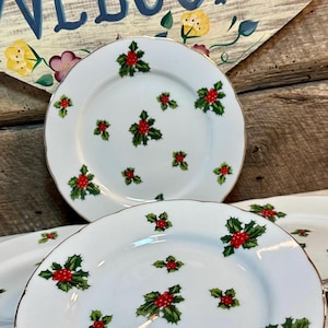 May include: Set of three white ceramic plates with a gold rim and a holly design. The plates are decorated with green holly leaves and red berries.