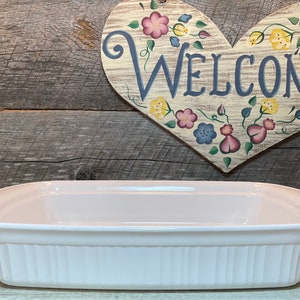 Puede incluir: Una fuente para hornear blanca, de forma ovalada, con laterales acanalados verticales. La fuente tiene un interior liso y un borde ligeramente elevado. En el fondo, un letrero de madera en forma de corazón con la palabra "WELCOME" y detalles florales.