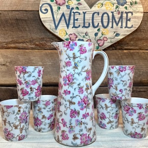 May include: A set of seven white ceramic cups and a pitcher with a floral pattern in shades of pink, blue, and gray. The cups and pitcher have a gold trim around the rim.