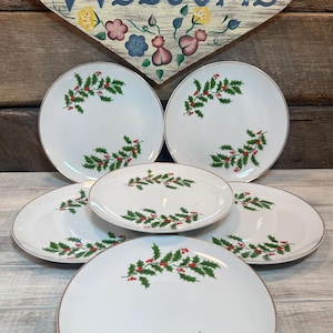 May include: A set of six white ceramic plates with a gold rim. Each plate features a green holly and red berry design. The plates are arranged on a wooden surface, with a heart-shaped sign that says "WELCOME" in the background.