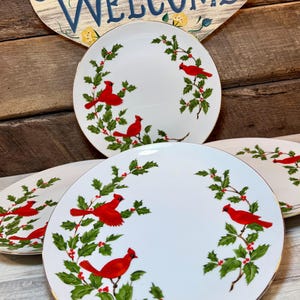 May include: A set of white ceramic plates featuring a festive pattern of bright red cardinal birds perched on green holly branches with red berries. Each plate has a thin gold rim, and the design is arranged in a circular wreath-like motif.