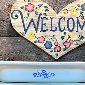 以下が含まれることがあります： 青い花柄の白い長方形のベーキングディッシュ。ディッシュは、青い文字で「WELCOME」と書かれたハート型の看板のある木製の表面の上に置かれています。