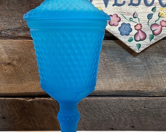 May include: A vintage, turquoise glass candy dish with a lid. The dish features a diamond pattern and a pedestal base. The lid is topped with a pointed finial. The background includes a wooden surface and a welcome sign.