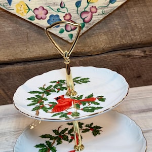 May include: A two-tier serving stand featuring white porcelain plates with scalloped edges and gold trim. Each plate is decorated with a red cardinal bird perched on green holly branches with red berries. A gold-tone metal handle connects the tiers.