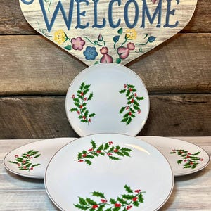 May include: Four white ceramic plates with a gold rim and a holly berry design. The holly design features green leaves and red berries. The plates are arranged in a group, with a wooden sign that says "WELCOME" in the background.