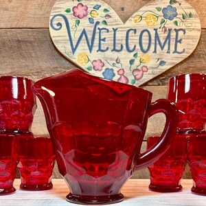 Puede incluir: Una jarra de vidrio rojo rubí con un juego a juego de seis vasos. La jarra y los vasos tienen un diseño geométrico texturizado. Un letrero de madera en forma de corazón con la palabra "Welcome" está en el fondo.