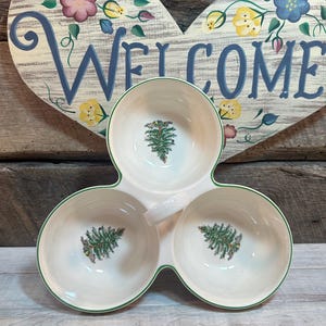 Puede incluir: Un plato blanco de tres secciones con ribete verde y asa. Cada sección presenta un diseño de pino verde y marrón. El plato está frente a un letrero de bienvenida de madera con detalles florales.