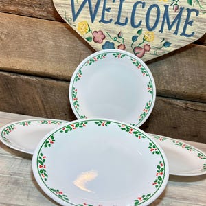 Puede incluir: Cuatro platos blancos con un borde verde y rojo de bayas de acebo. Los platos están dispuestos sobre una superficie de madera clara. Un letrero de madera con la palabra "WELCOME" está en el fondo.