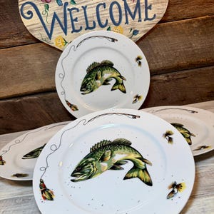 May include: A set of white ceramic plates featuring a green and yellow fish illustration in the center, surrounded by a thin black line depicting a fishing line with colorful lures. The plates have a speckled finish and a smooth, round edge.