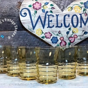 Puede incluir: Seis vasos de vidrio ámbar con un diseño acanalado. Los vasos están dispuestos en una fila sobre una superficie de madera.