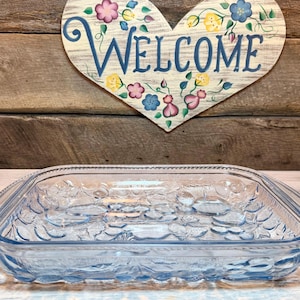 May include: A light blue rectangular glass baking dish with floral embossed detailing. The dish is sitting in front of a wooden heart-shaped sign that says "WELCOME" in blue lettering, with floral accents.