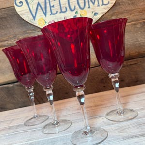 May include: Four ruby red wine glasses with clear stems and bases. The glasses have a tulip-shaped bowl. The background includes a wooden sign with the word "WELCOME" in blue script.