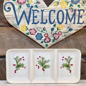 May include: A heart-shaped wooden sign with a white wood grain finish, painted with blue text that reads WELCOME and decorated with colorful flowers. Below it is a white rectangular ceramic serving tray with three sections and holly.