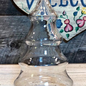 Pode incluir: Um frasco de vidro transparente em forma de árvore de Natal, com uma base larga e uma parte superior pontiaguda. O frasco está vazio e tem um leve brilho iridescente.