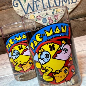 May include: Two clear drinking glasses featuring the Pac-Man video game characters. The glasses have a blue, yellow, red, and black design. The background includes a wooden heart-shaped sign that says "WELCOME".