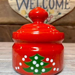 Könnte beinhalten: Kleines, rotes Keramikgefäß mit Deckel, mit einem grünen Weihnachtsbaumdesign mit weißen Punkten. Das Gefäß ist glänzend und scheint ein dekorativer Gegenstand zu sein. Das Wort "WELCOME" ist im Hintergrund sichtbar.