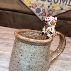 May include: A speckled ceramic mug with a whimsical pig figurine perched on the handle. The pig is white with brown spots and pink details. The mug has a rustic, handmade appearance.
