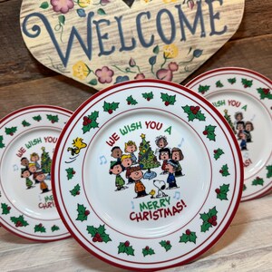May include: Three white ceramic plates with a red rim and holly berry design. The plates feature a cartoon image of children singing around a Christmas tree, with the text "WE WISH YOU A MERRY CHRISTMAS!"
