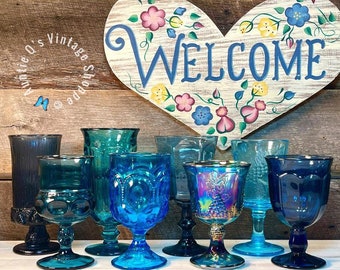 Könnte beinhalten: Eine Sammlung von Vintage-Bechern und Gläsern aus blauem Glas, einige mit strukturiertem Design, auf einer Holzoberfläche angeordnet mit einem bemalten Herzschild, auf dem "WELCOME" steht.