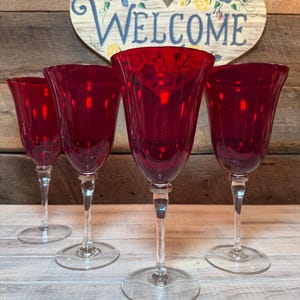 May include: Four ruby red wine glasses with clear stems and bases. The glasses have a tulip-shaped bowl and are arranged in a row. A wooden heart-shaped sign with the word "Welcome" is in the background.