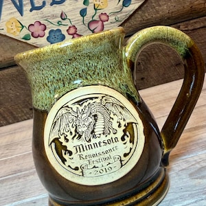 May include: A ceramic mug with a two-tone brown and green glaze featuring a circular medallion on the front. The medallion depicts a dragon and the text Minnesota Renaissance Festival 2019. The mug has a sturdy handle and a wide, flared rim.