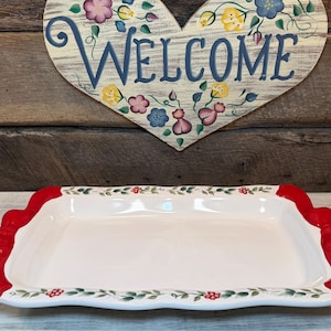 May include: A rectangular white ceramic serving tray featuring bright red handles on each side. The rim is decorated with a painted pattern of green leaves and small red berries. The tray has a smooth, glossy finish and a slightly scalloped edge.
