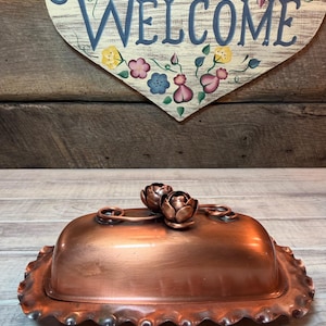 May include: A copper butter dish with a domed lid and a ruffled edge. Two decorative flower-shaped handles are on top. The background includes a wooden heart-shaped sign with the word "Welcome" and floral accents.